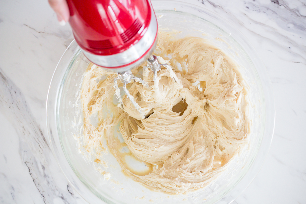 Softened cream cheese being stirred with shredded cheddar and sliced scallions in a glass mixing bowl on a kitchen counter