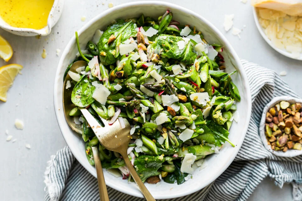 Spring salad ingredients arranged on a kitchen counter including asparagus, snap peas, radishes, lemons, mixed greens, fresh herbs, feta, almonds, olive oil, and Dijon mustard