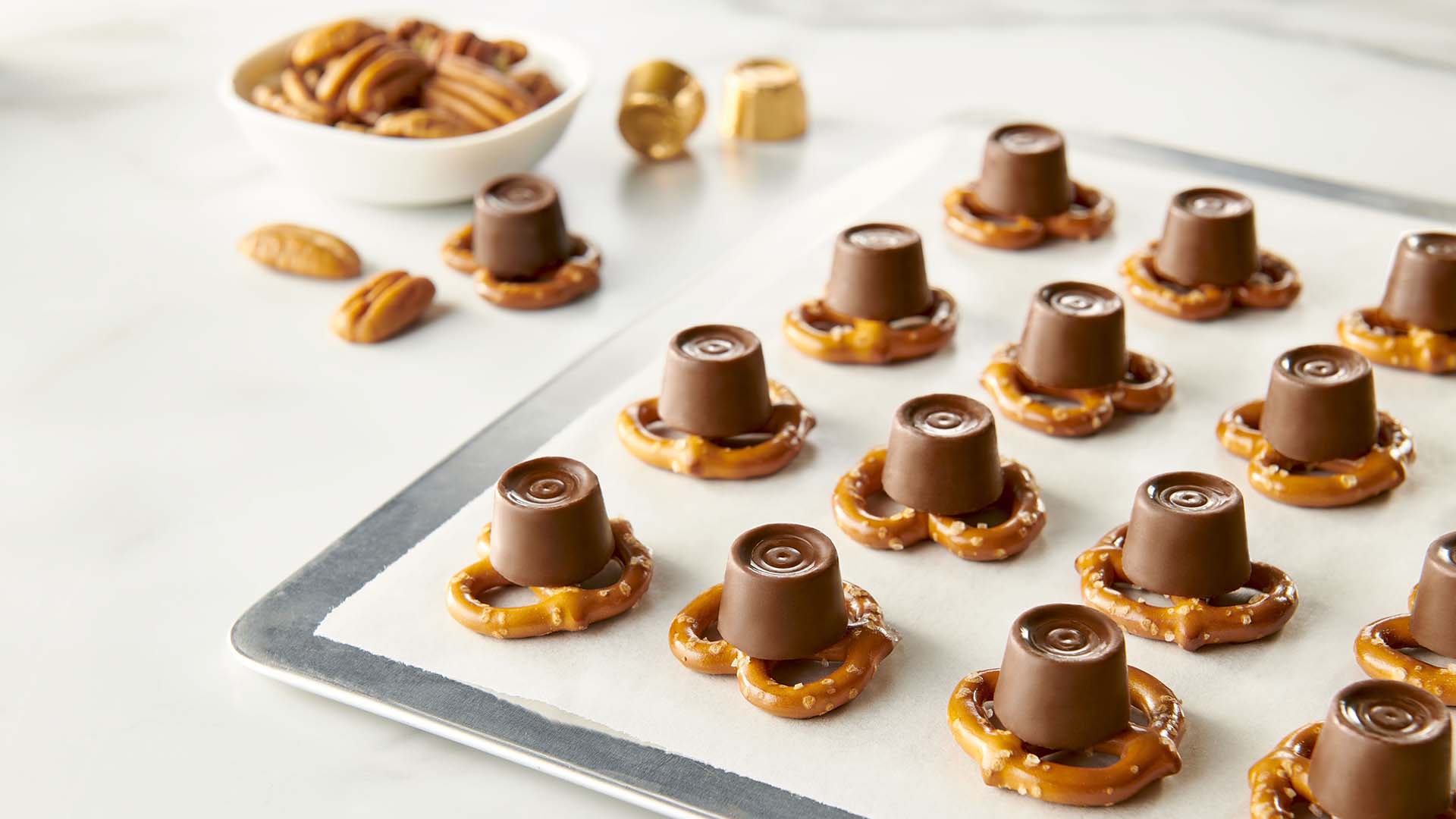 Square pretzels, unwrapped Rolo candies, and pecan halves arranged in small bowls on a clean kitchen counter