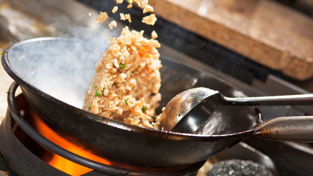 Stir-fried rice being tossed in a hot wok with a metal spatula, visible steam and browned rice grains
