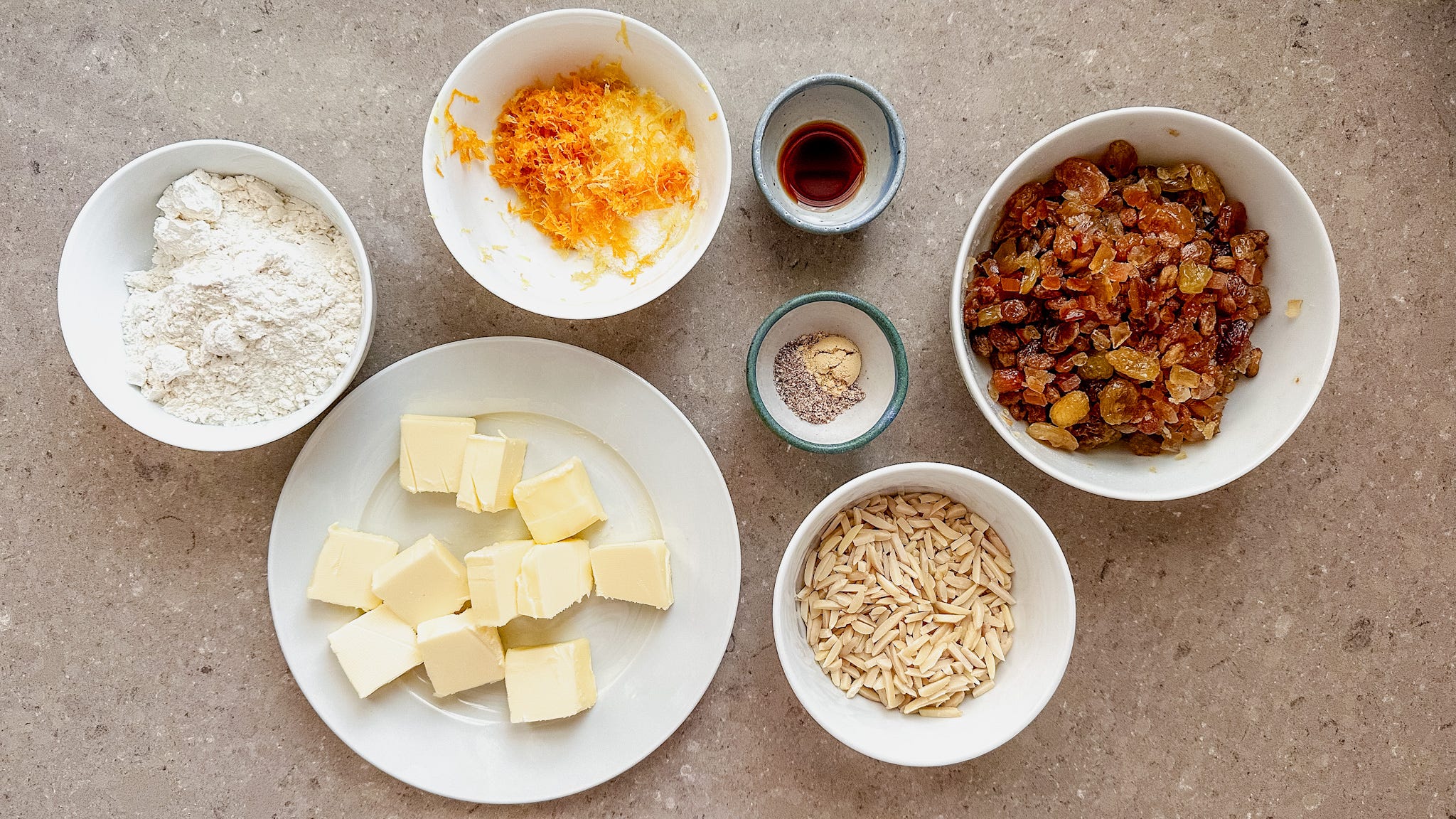 Stollen ingredients arranged on a light kitchen counter, including flour, butter, eggs, milk, yeast, dried fruit, candied citrus peel, almonds, and a small bowl of spices
