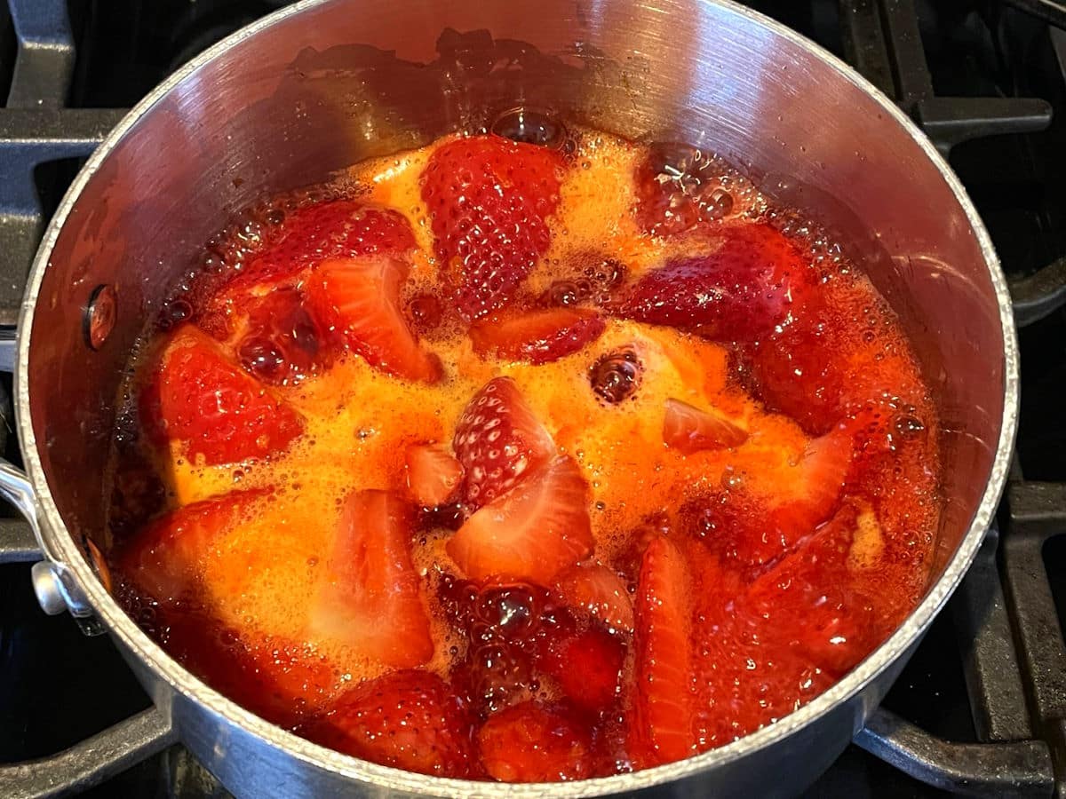 Strawberries simmering with sugar in a small saucepan on a stove, bubbling gently as they break down into a bright red syrup