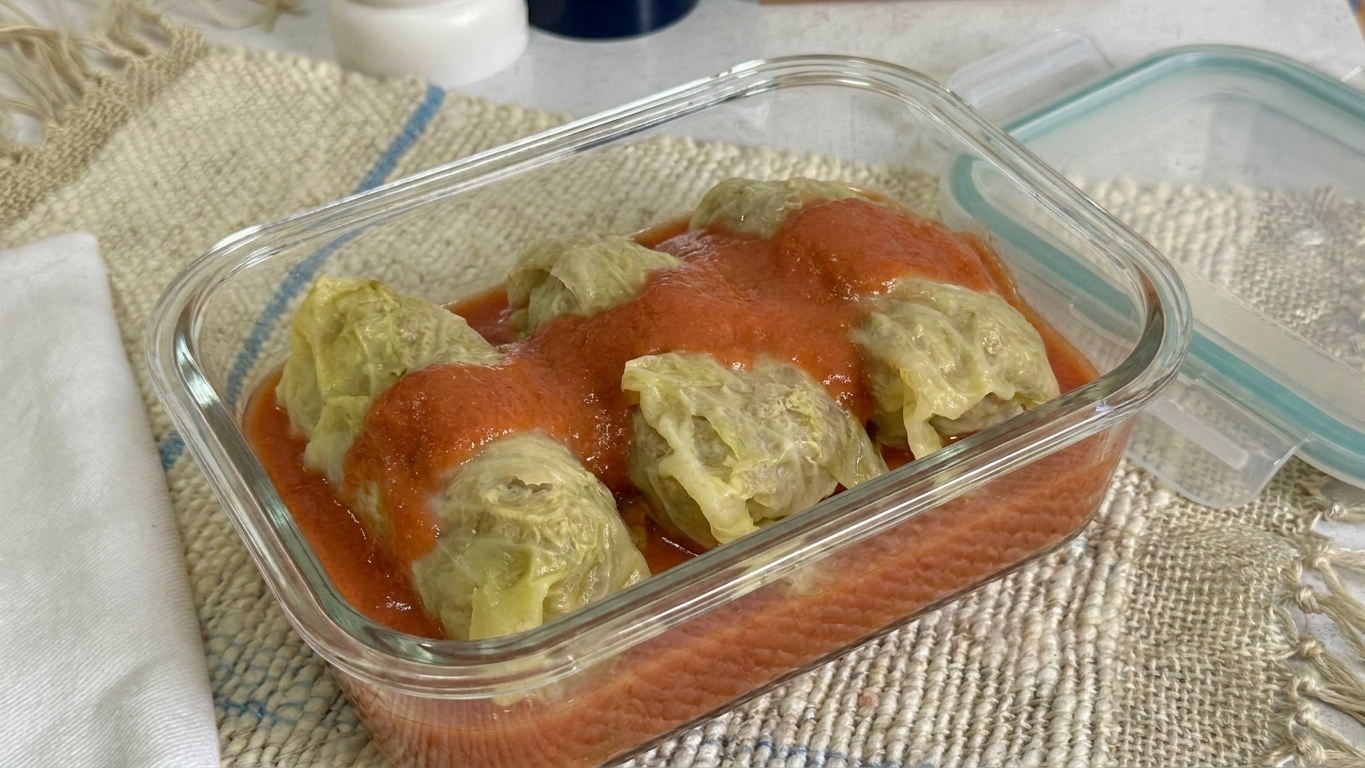 Stuffed cabbage rolls in a glass meal prep container with tomato sauce, lid set nearby on a kitchen counter