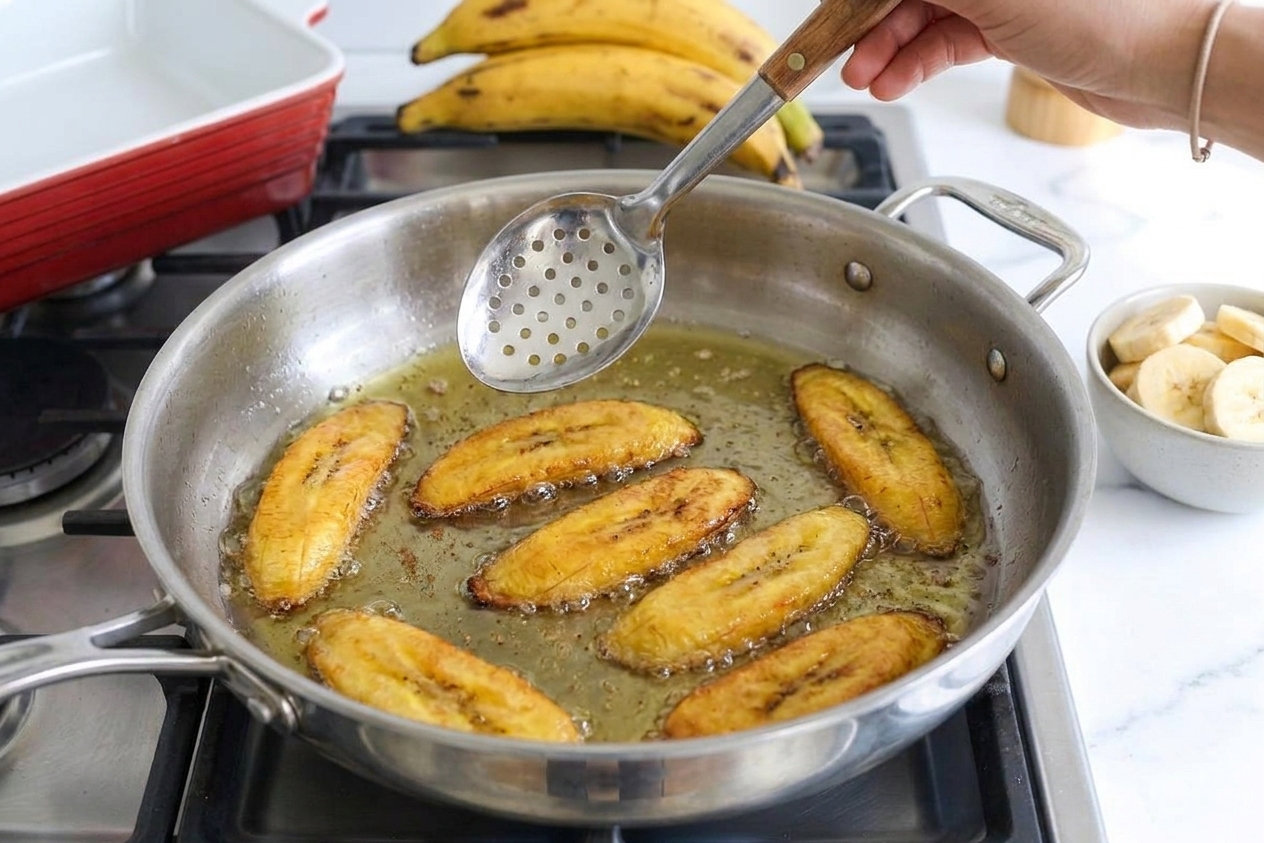 Sweet plantain slices frying in hot oil in a skillet with a slotted spoon hovering above them