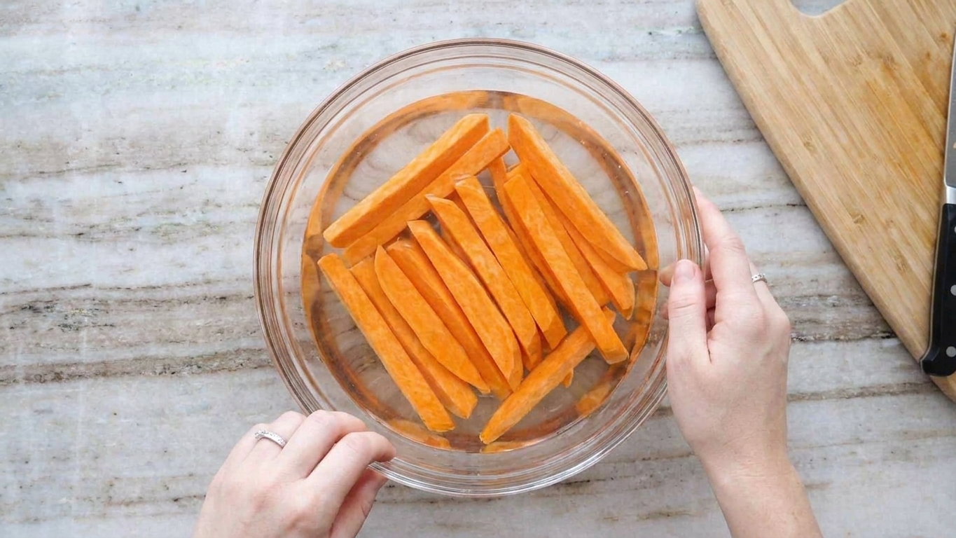 Sweet potato sticks soaking in a clear glass bowl of water on a kitchen counter