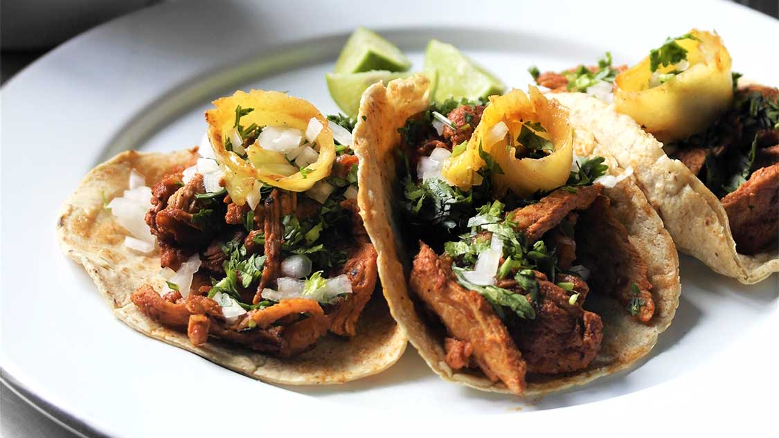 Tacos al pastor on a warm ceramic plate with charred pineapple, chopped onion and cilantro, lime wedges, and a small bowl of salsa on a wooden table in soft window light