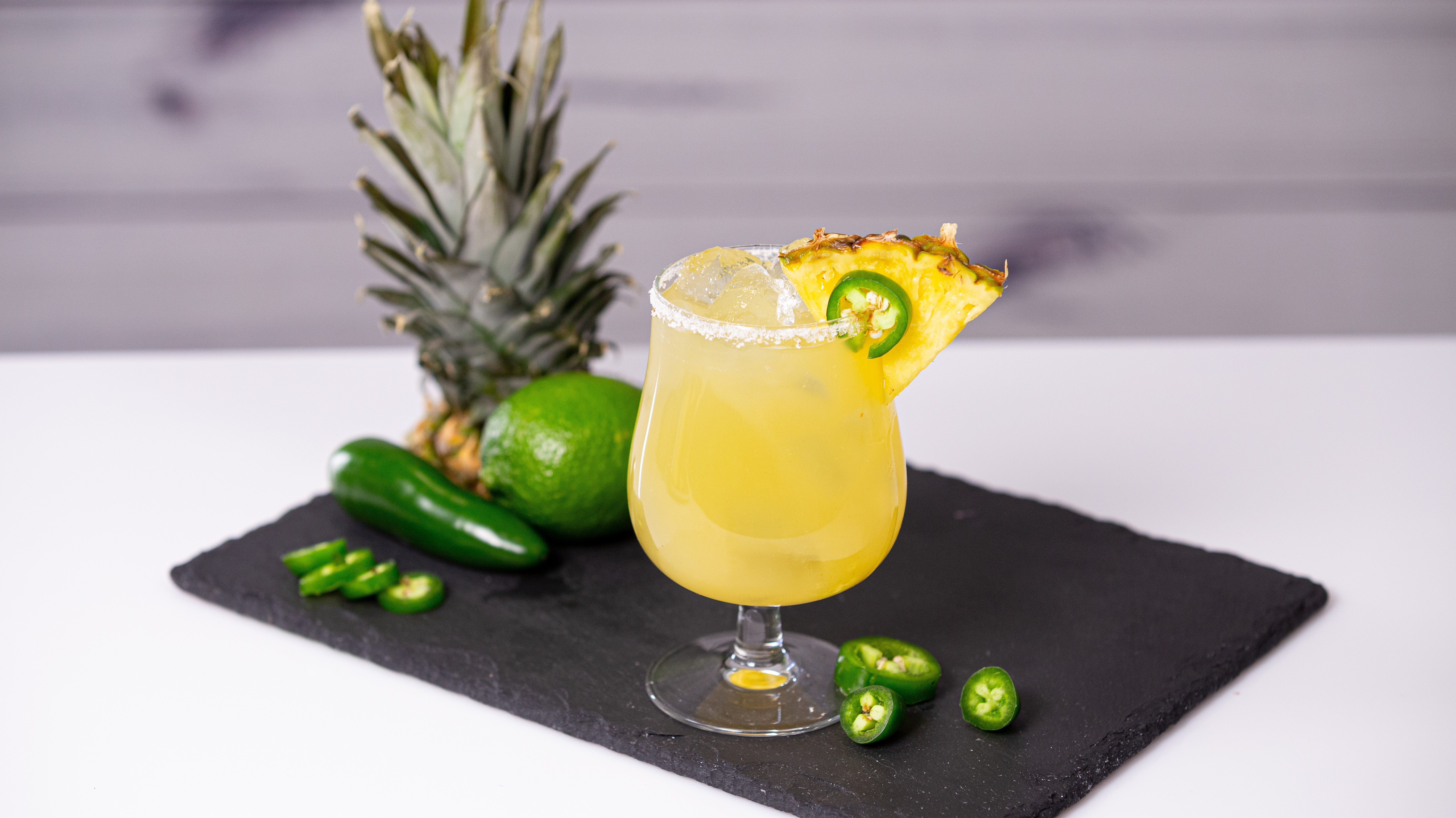 Tequila, pineapple juice, fresh limes, a sliced jalapeño, and a small dish of tajín and salt arranged on a kitchen counter, ready for cocktail prep