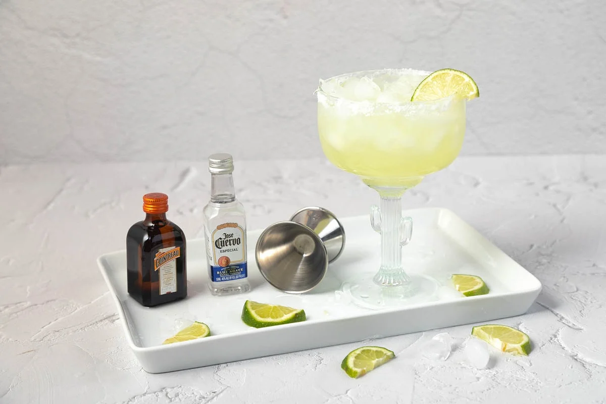 Tequila, triple sec, fresh limes, a small bowl of salt, and ice arranged on a bright kitchen counter ready for making margaritas