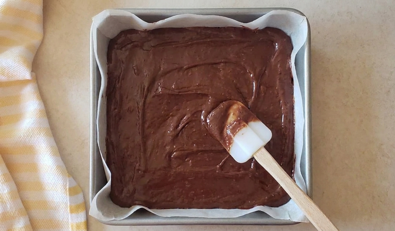 Thick brownie batter spread evenly in a parchment lined square baking pan