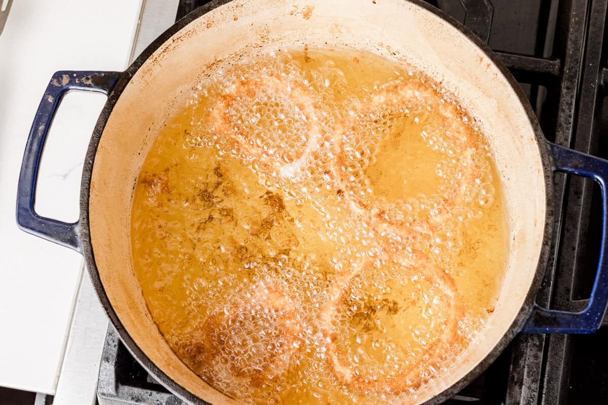Thick onion rings frying in bubbling oil inside a Dutch oven on a stovetop while a spider strainer lifts one ring above the oil