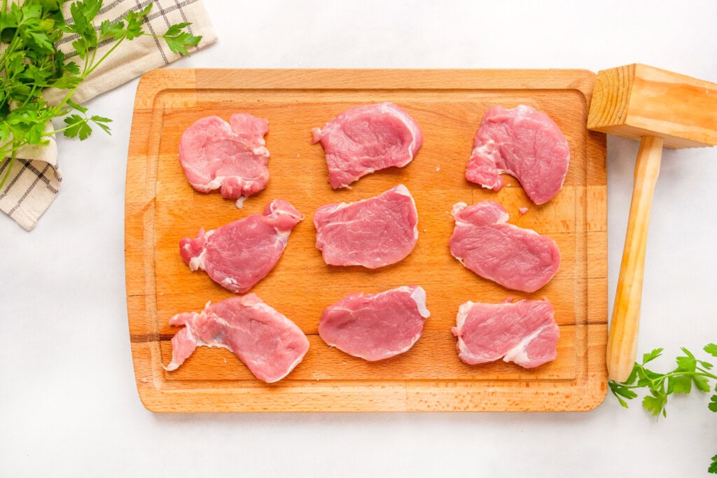 Thick slices of raw pork tenderloin cut into medallions on a wooden cutting board with a chef's knife and small bowls of salt and spices nearby