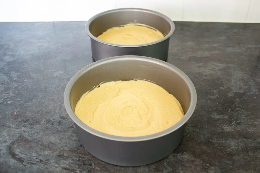 Thick vanilla cake batter being smoothed with a spatula in a round cake pan on a kitchen counter before baking