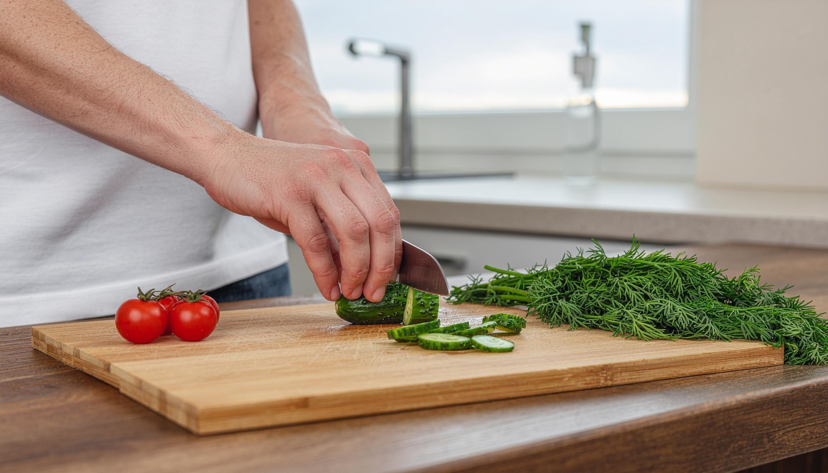 Thin cucumber rounds being sliced on a wooden cutting board with a chef's knife in a bright home kitchen