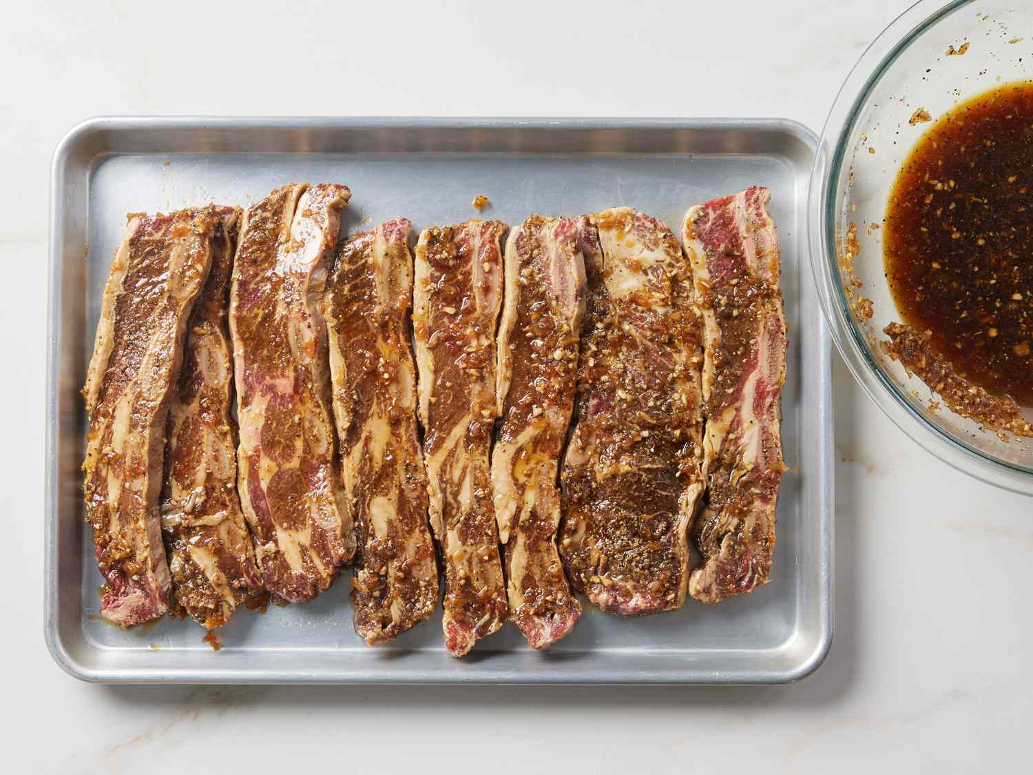 Thin-cut marinated LA-style short ribs arranged on a foil-lined sheet pan under an oven broiler, edges beginning to caramelize