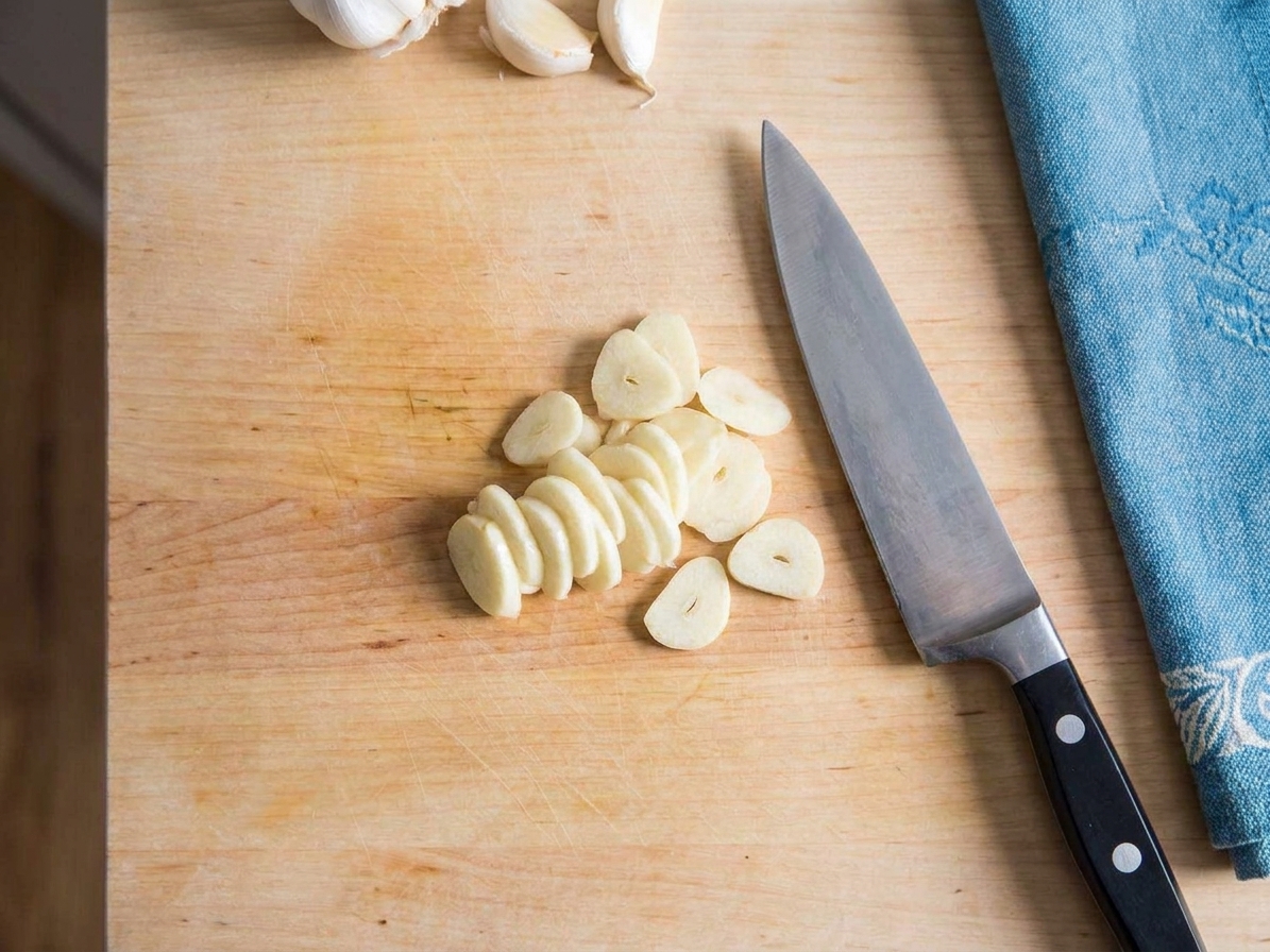 Thin garlic slices on a wooden cutting board next to a chef's knife