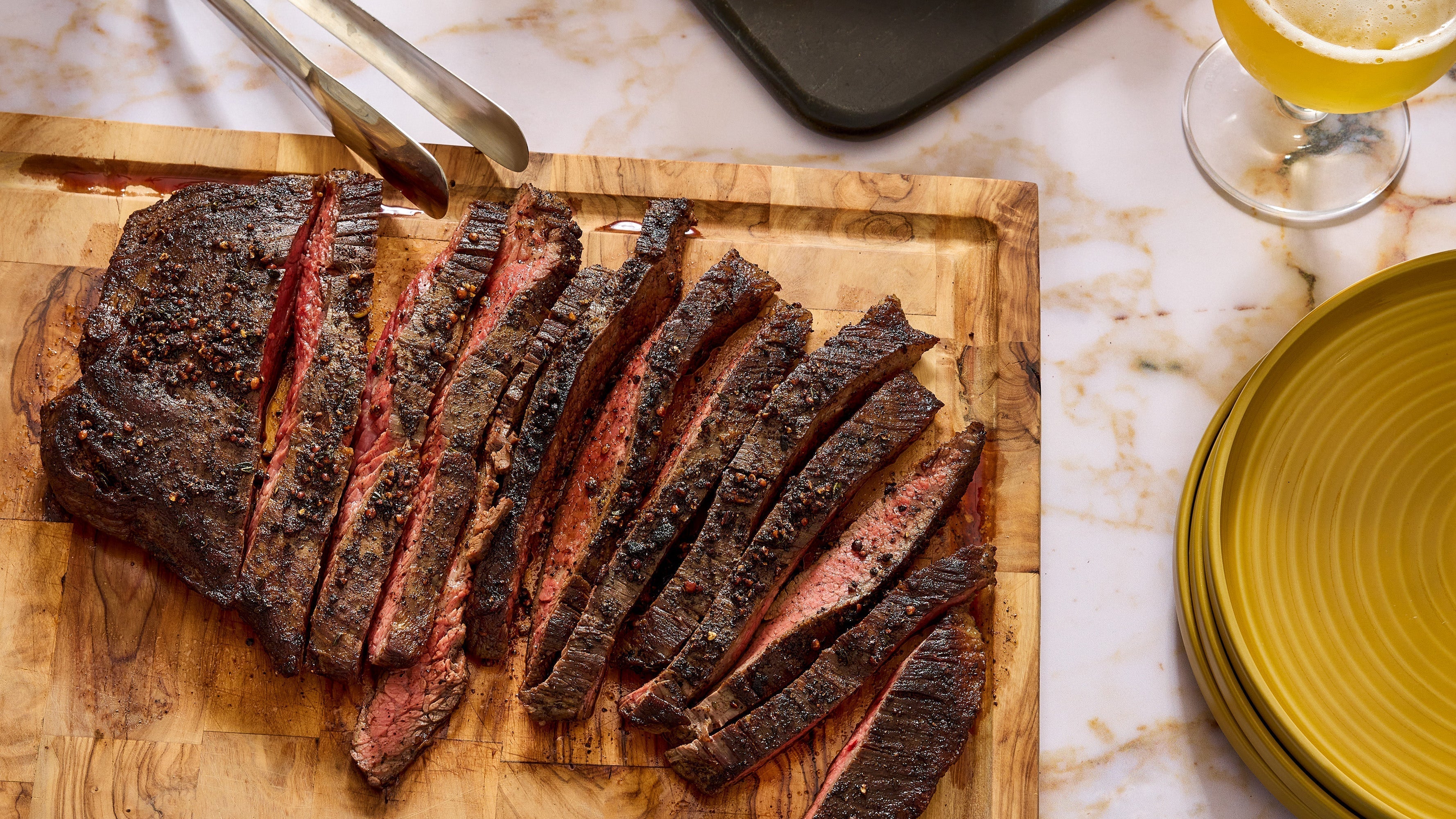 Thin slices of flank steak on a wooden cutting board with a chef's knife, ready for the slow cooker on a bright kitchen counter