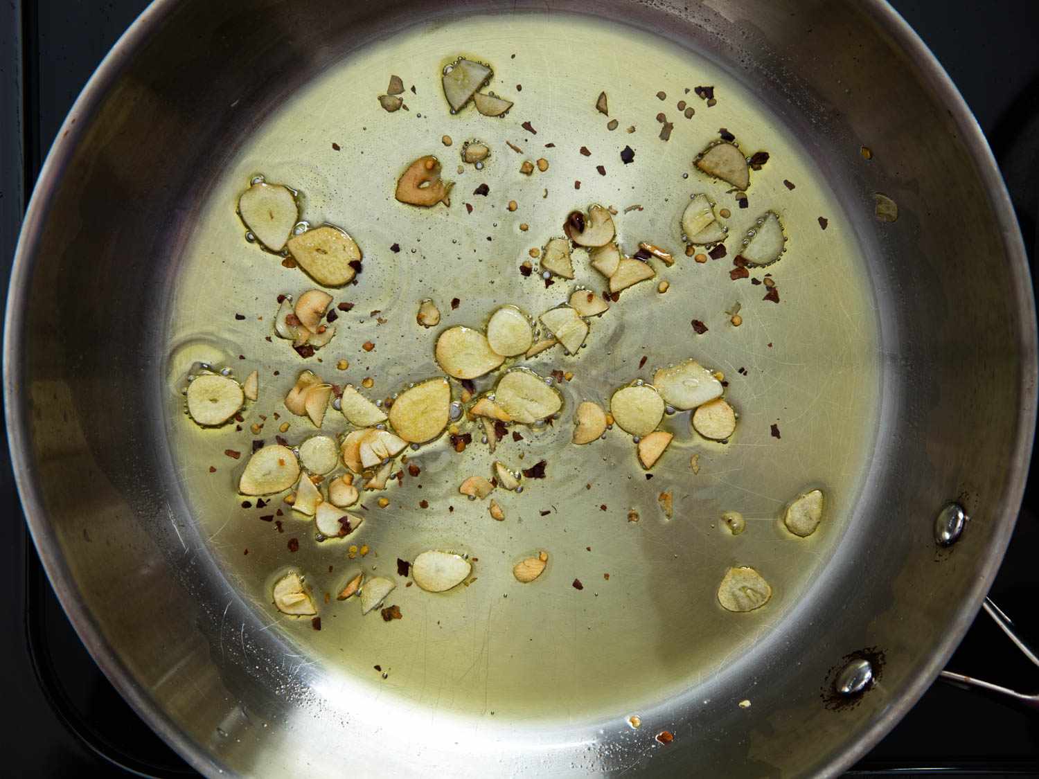 Thin slices of garlic sizzling in olive oil in a stainless steel skillet on a stovetop