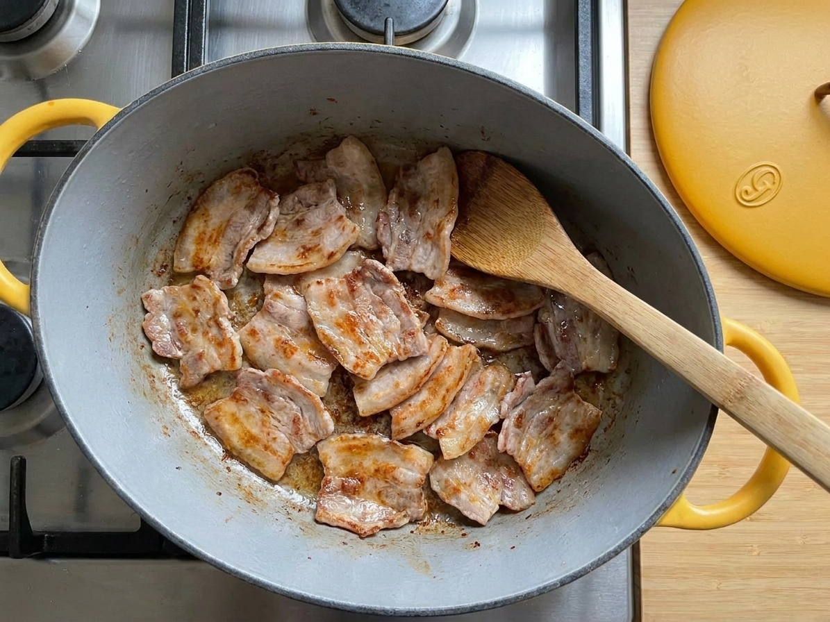 Thin slices of pork browning in a Dutch oven on a stovetop with a wooden spoon nearby