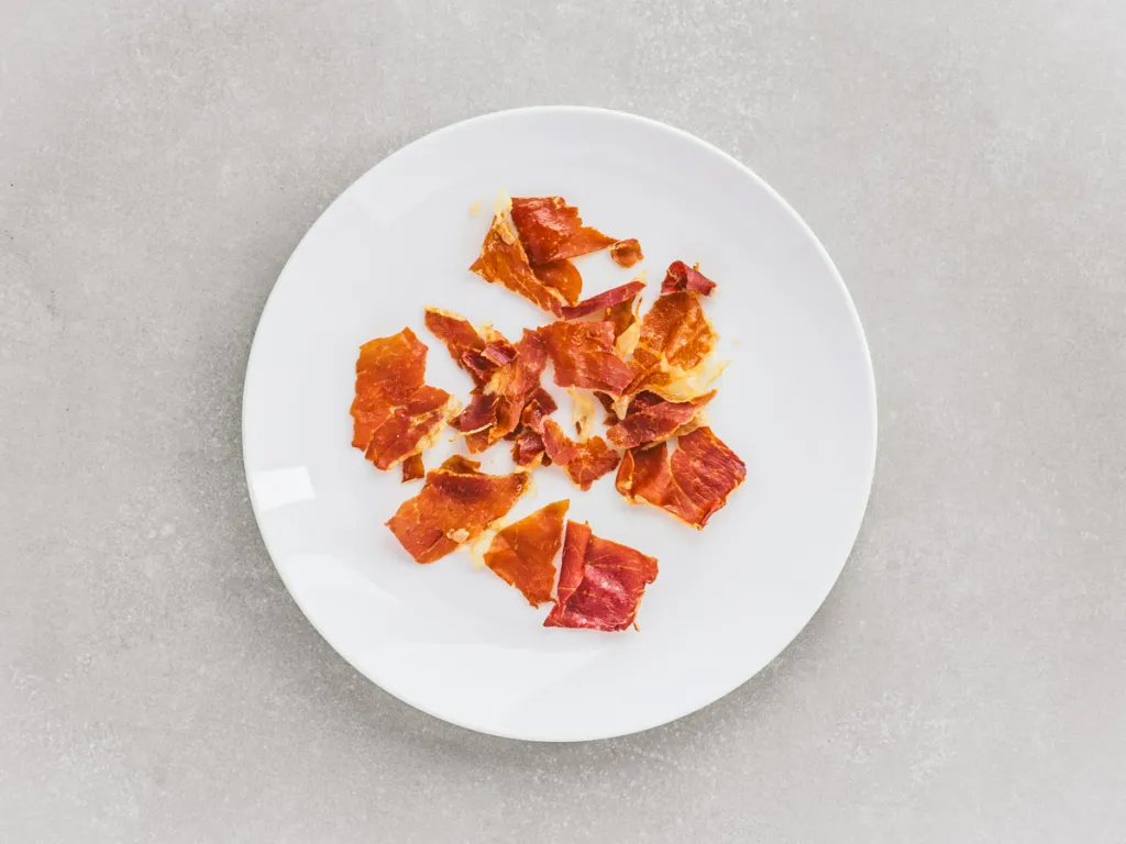 Thin slices of prosciutto sizzling in a skillet on a stovetop, edges curling as they turn crisp