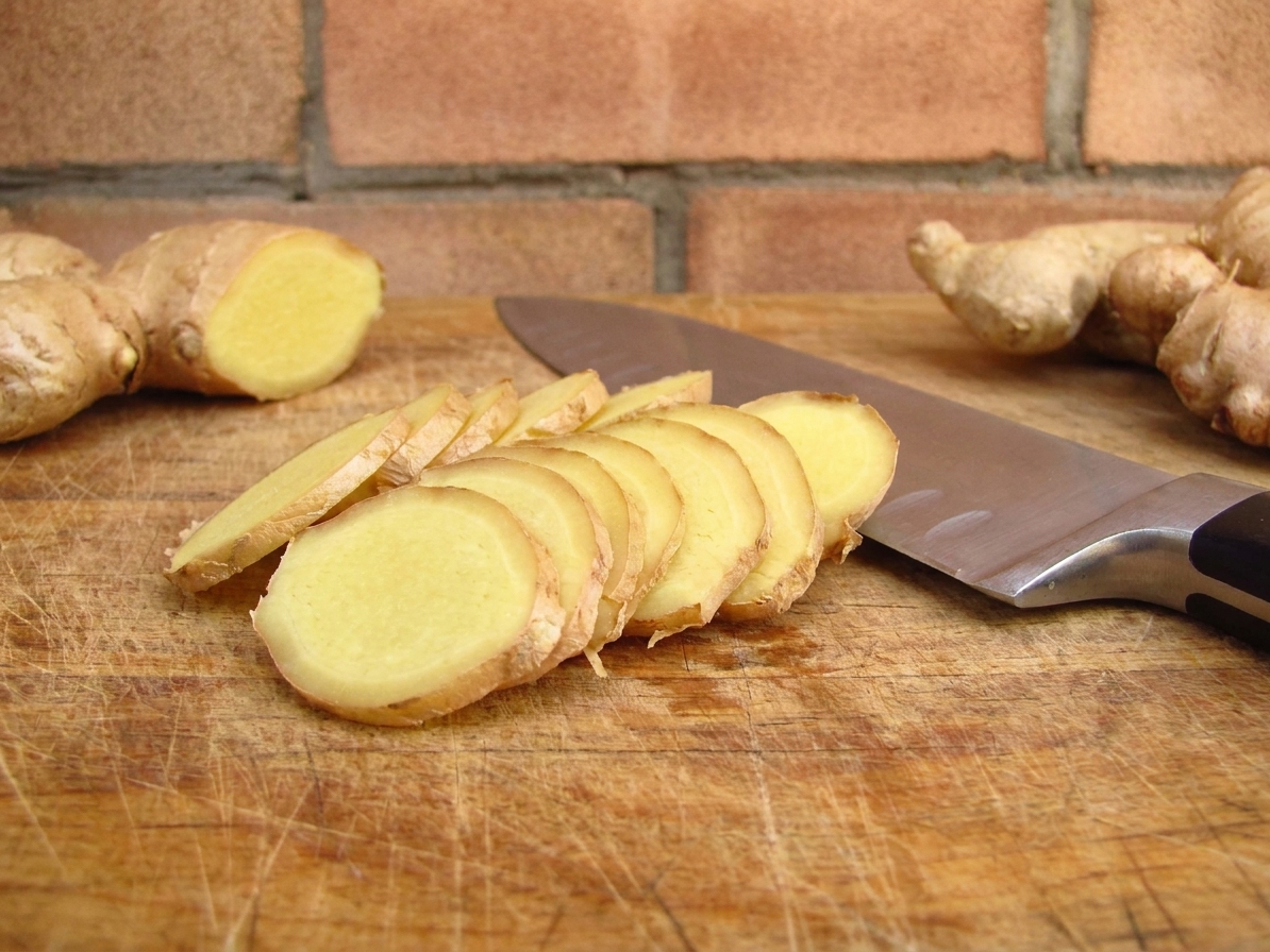 Thinly sliced fresh ginger on a wooden cutting board with a chef's knife beside it on a kitchen counter