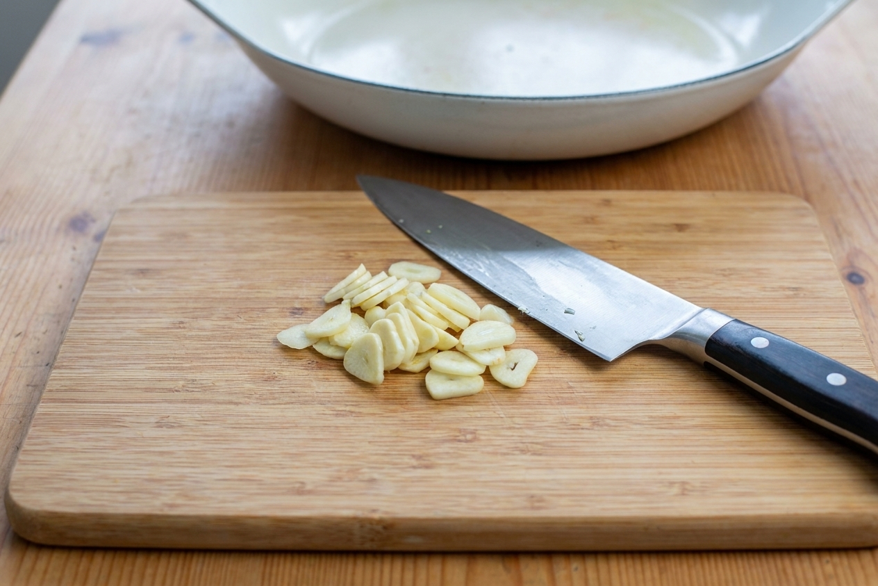Thinly sliced garlic cloves on a cutting board beside a chef's knife on a home kitchen counter
