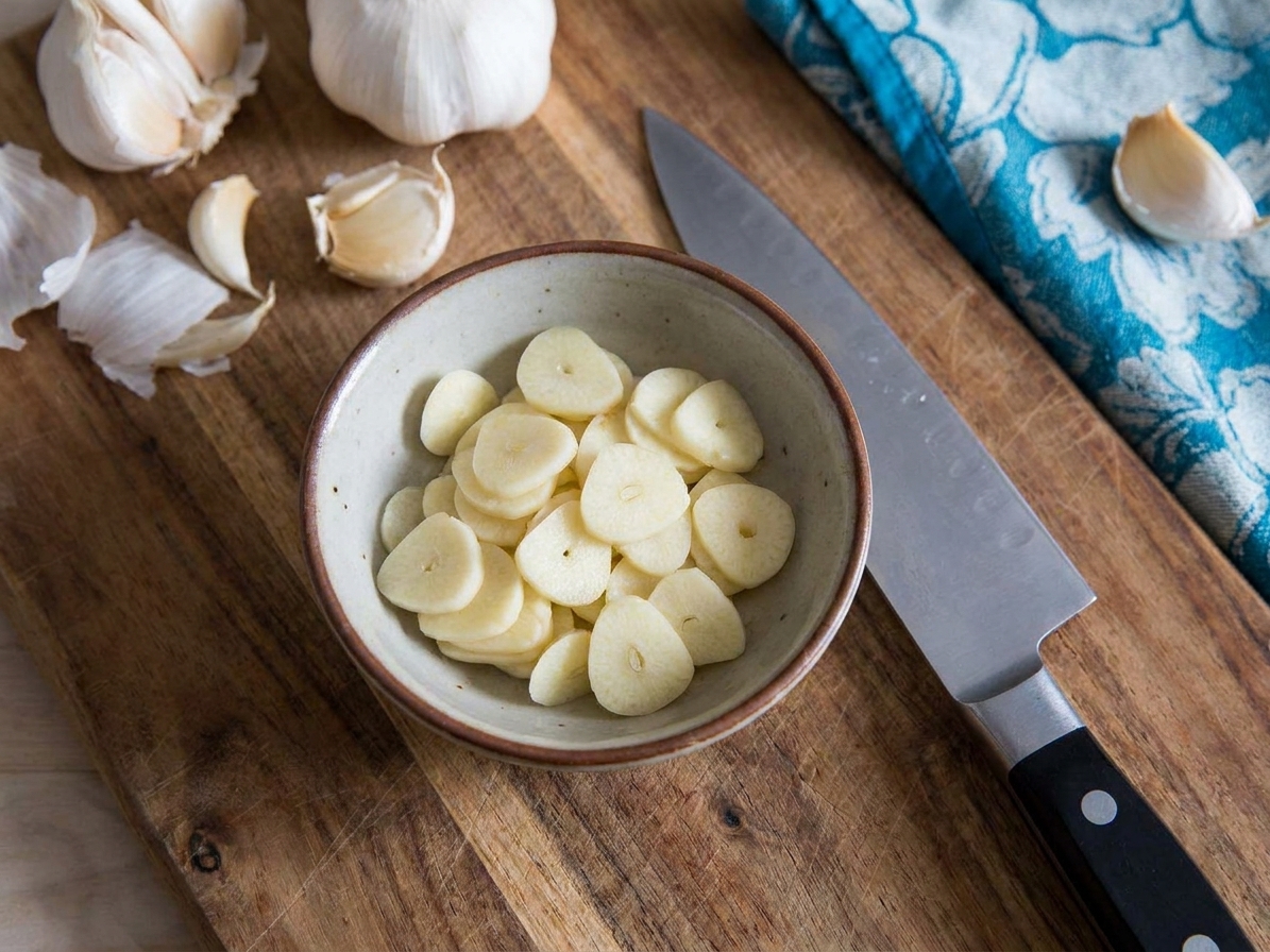 Thinly sliced garlic coins in a small ceramic bowl next to a chef's knife on a wooden cutting board