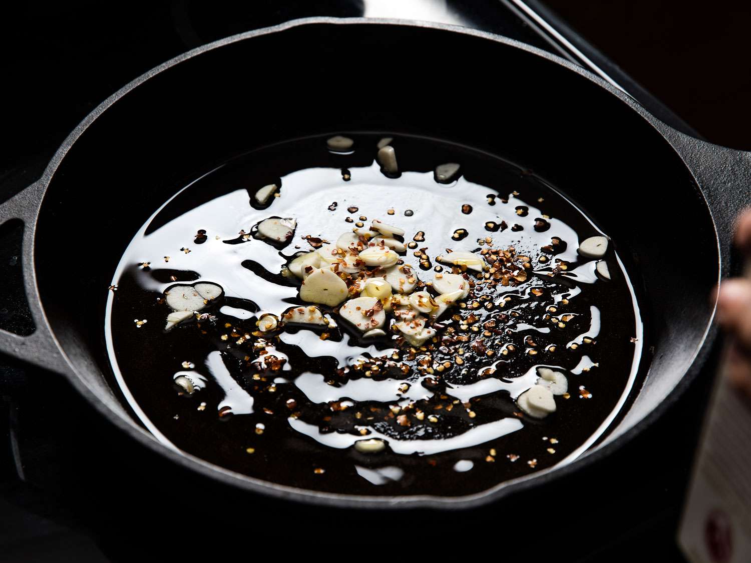 Thinly sliced garlic gently cooking in olive oil in a large skillet on a stove, with a wooden spoon nearby