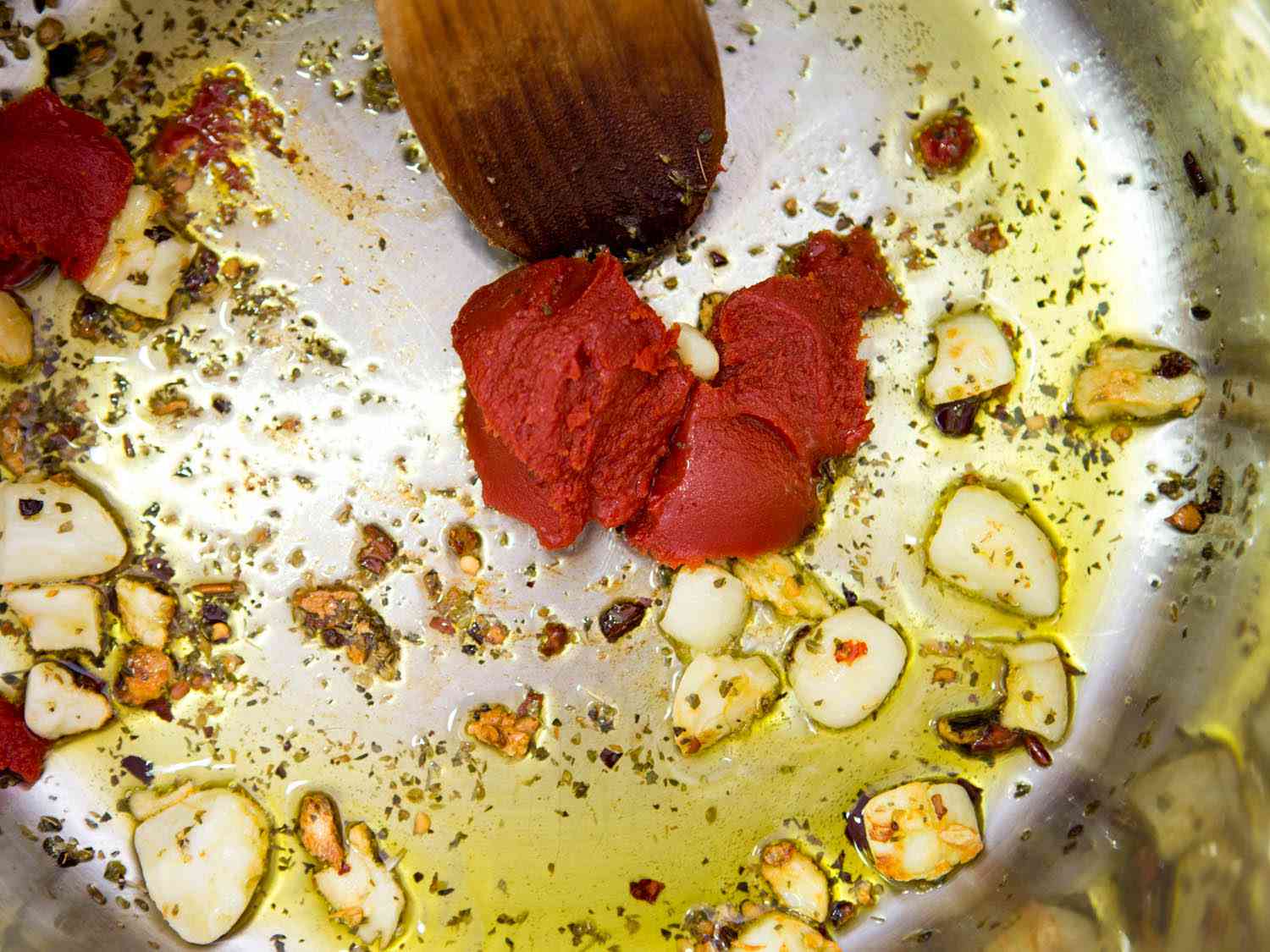 Thinly sliced garlic on a cutting board next to a small bowl of tomato paste and red pepper flakes on a home kitchen counter