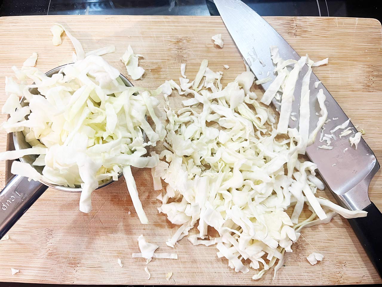 Thinly sliced green cabbage and onion piled on a wooden cutting board with a chef’s knife on a bright kitchen counter