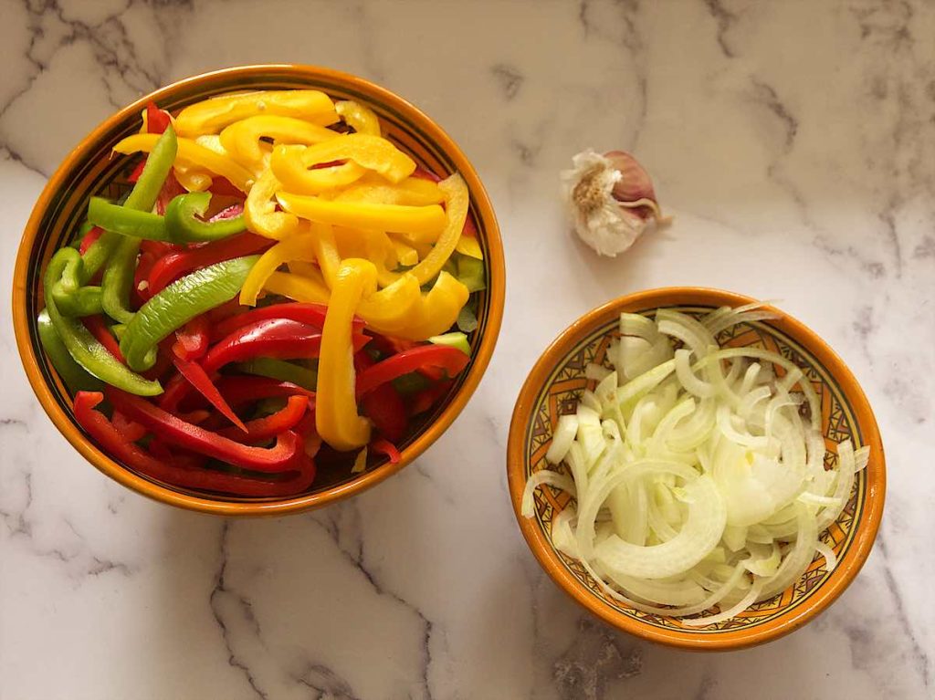 Thinly sliced onions, bell peppers, and garlic on a cutting board beside a skillet on a home kitchen counter