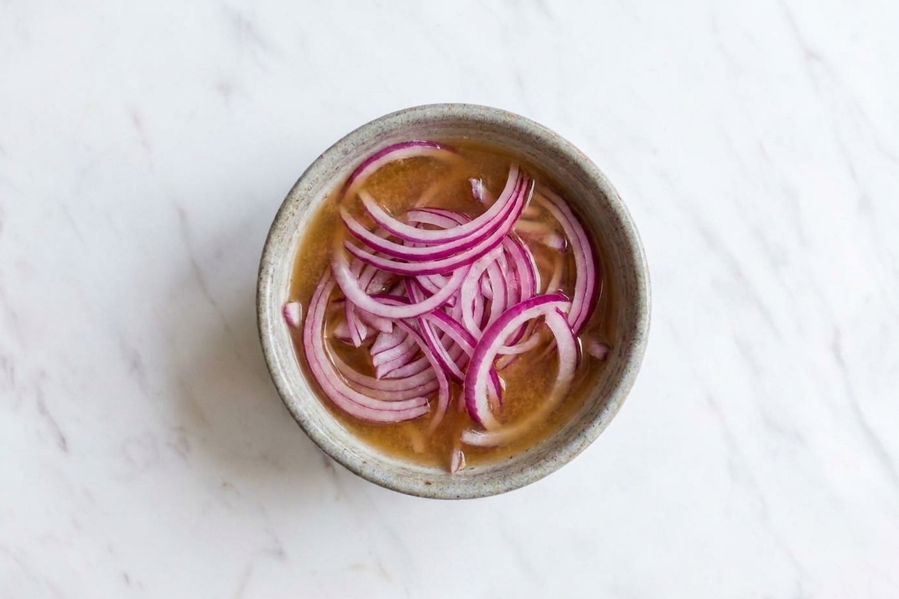 Thinly sliced red onions soaking in apple cider vinegar in a small glass bowl on a light kitchen counter