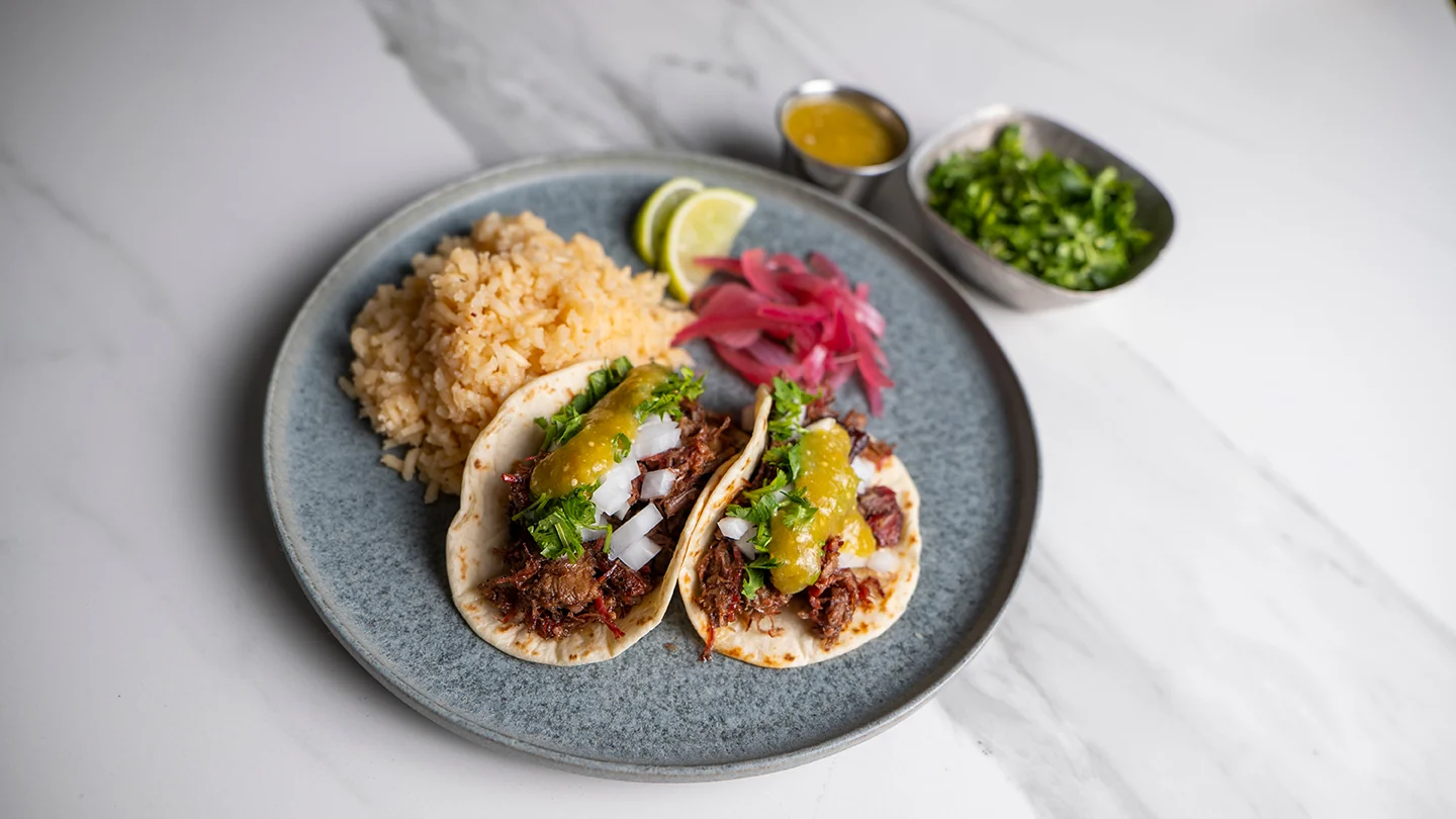 Three leftover brisket tacos on a stoneware plate with lime cilantro slaw and a drizzle of chipotle sauce on a wooden table