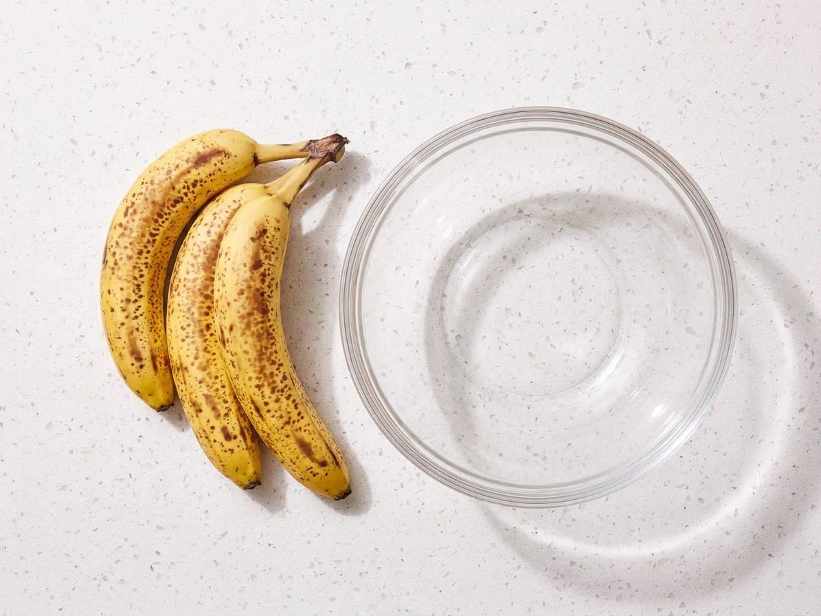 Three overripe bananas with brown speckles resting on a light-colored kitchen counter next to a mixing bowl