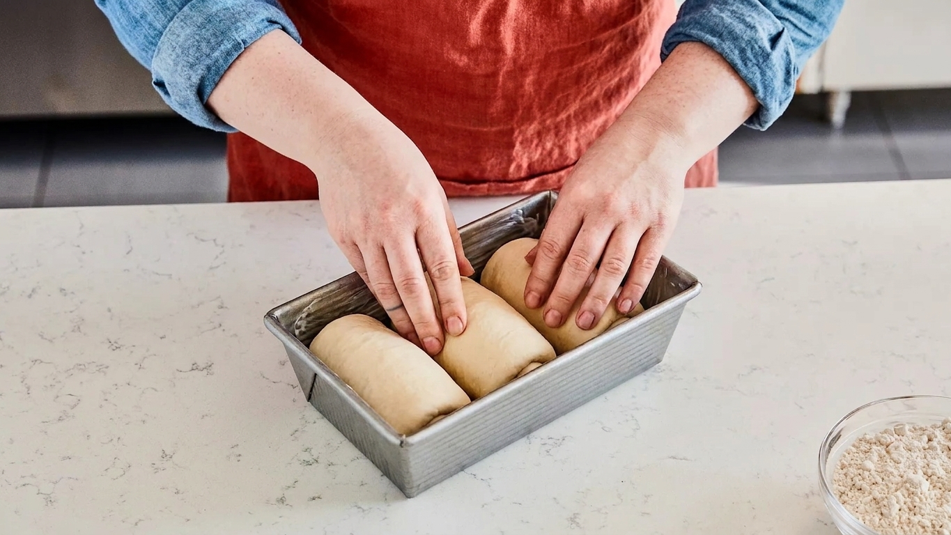 Three rolled dough logs being placed seam-side down into a greased loaf pan on a kitchen counter