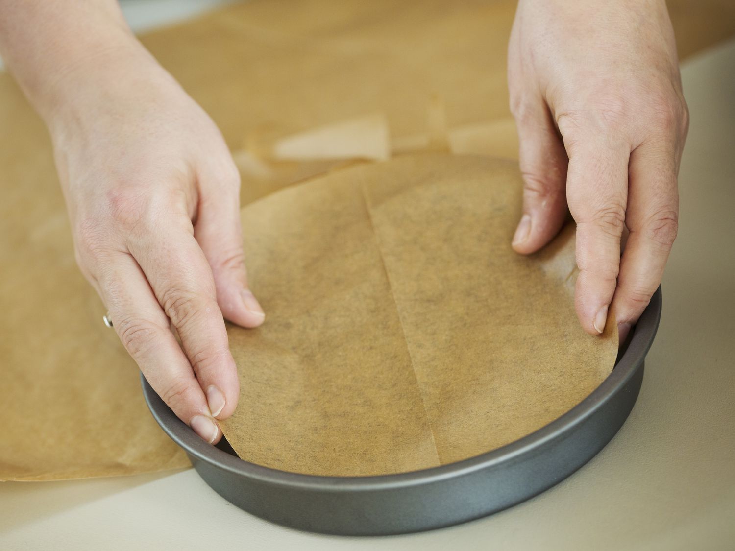 Three round cake pans lined with parchment paper circles on a kitchen counter next to a small bowl of butter for greasing
