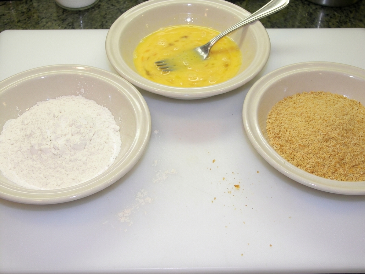 Three shallow bowls on a kitchen counter containing flour, beaten eggs, and fine breadcrumbs ready for breading cutlets