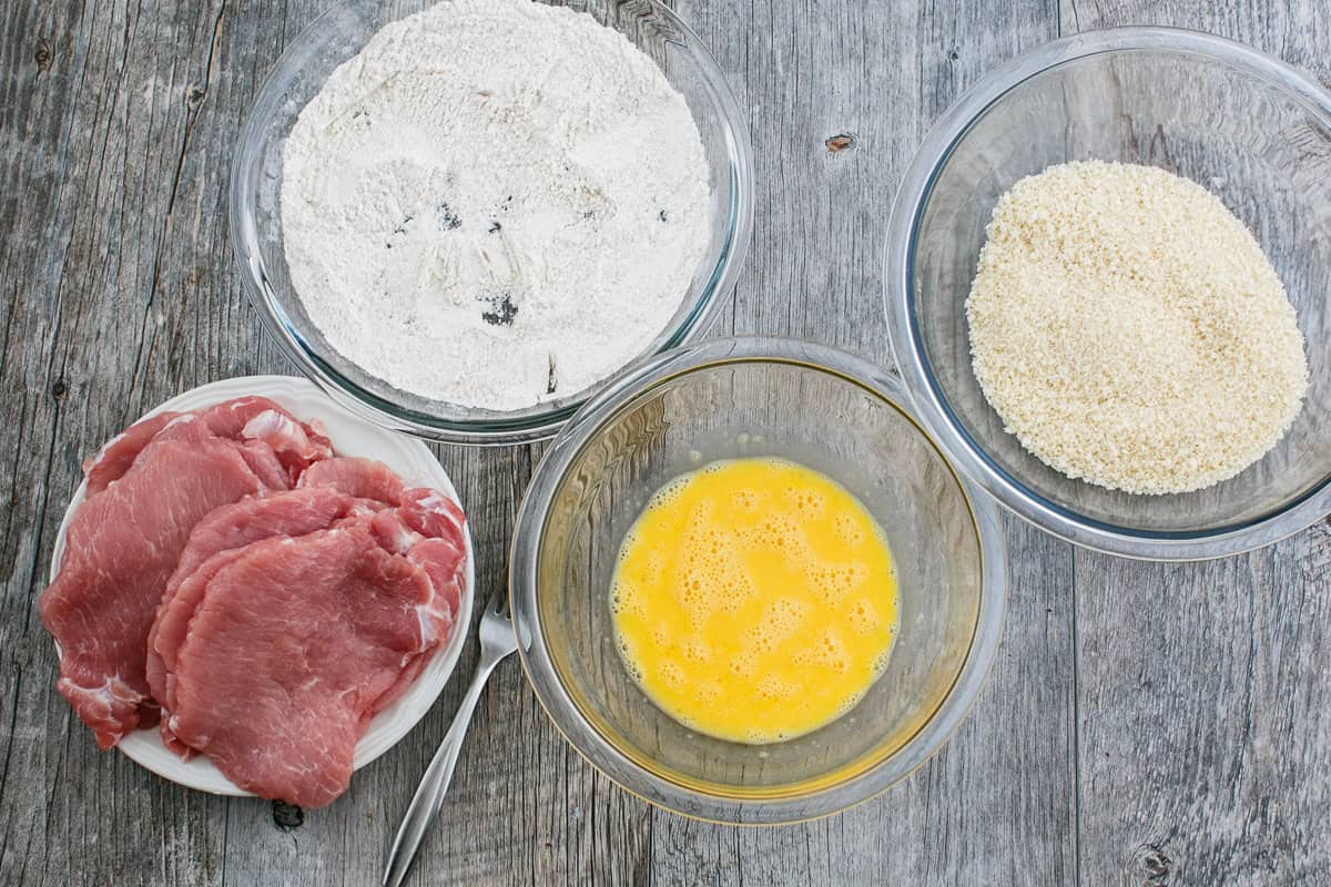 Three shallow bowls set up as a breading station with flour, beaten eggs, and panko breadcrumbs on a kitchen counter