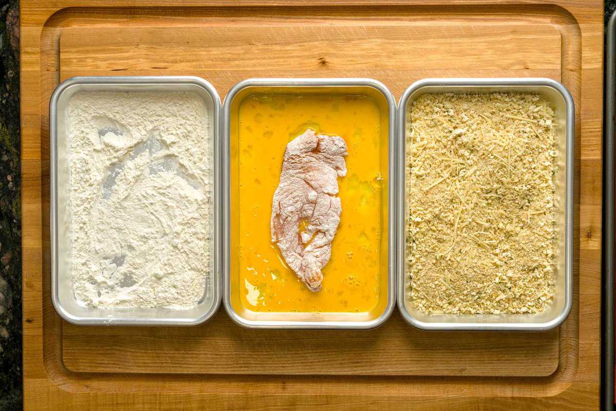 Three shallow bowls set up for breading with flour, beaten eggs, and a breadcrumb-parmesan mixture on a kitchen counter with chicken cutlets nearby