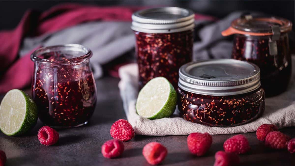 Three small jars filled with raspberry jam cooling on a kitchen towel on the counter, lids set nearby