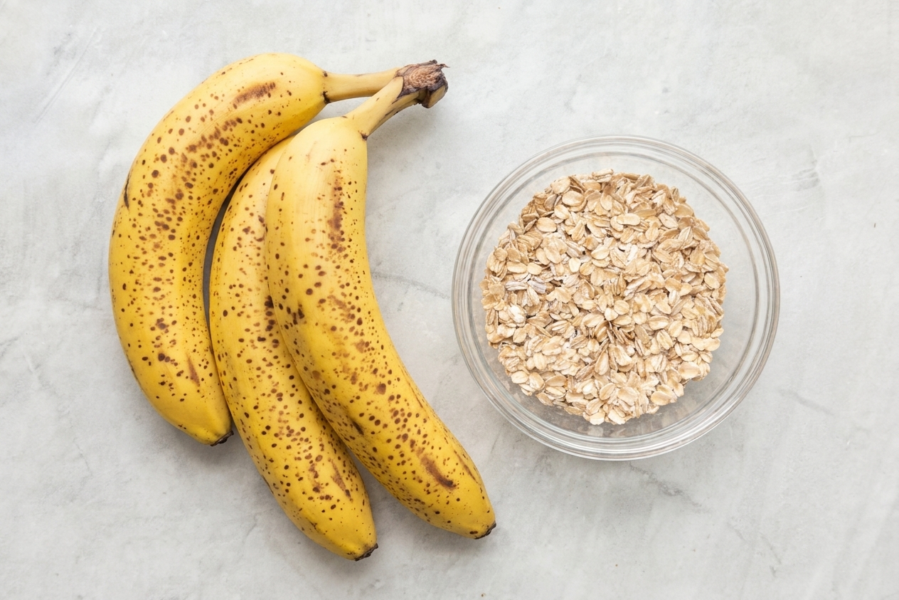 Three very ripe bananas with brown speckles resting on a light colored kitchen counter next to a small bowl of oats