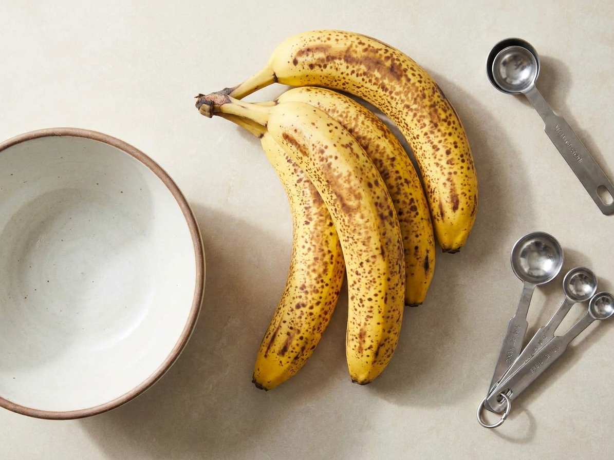 Three very ripe, speckled bananas resting on a light stone kitchen counter next to a mixing bowl and measuring spoons in natural light