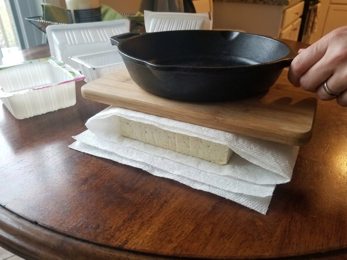 Tofu block wrapped in paper towels with a cutting board and a heavy skillet on top on a kitchen counter