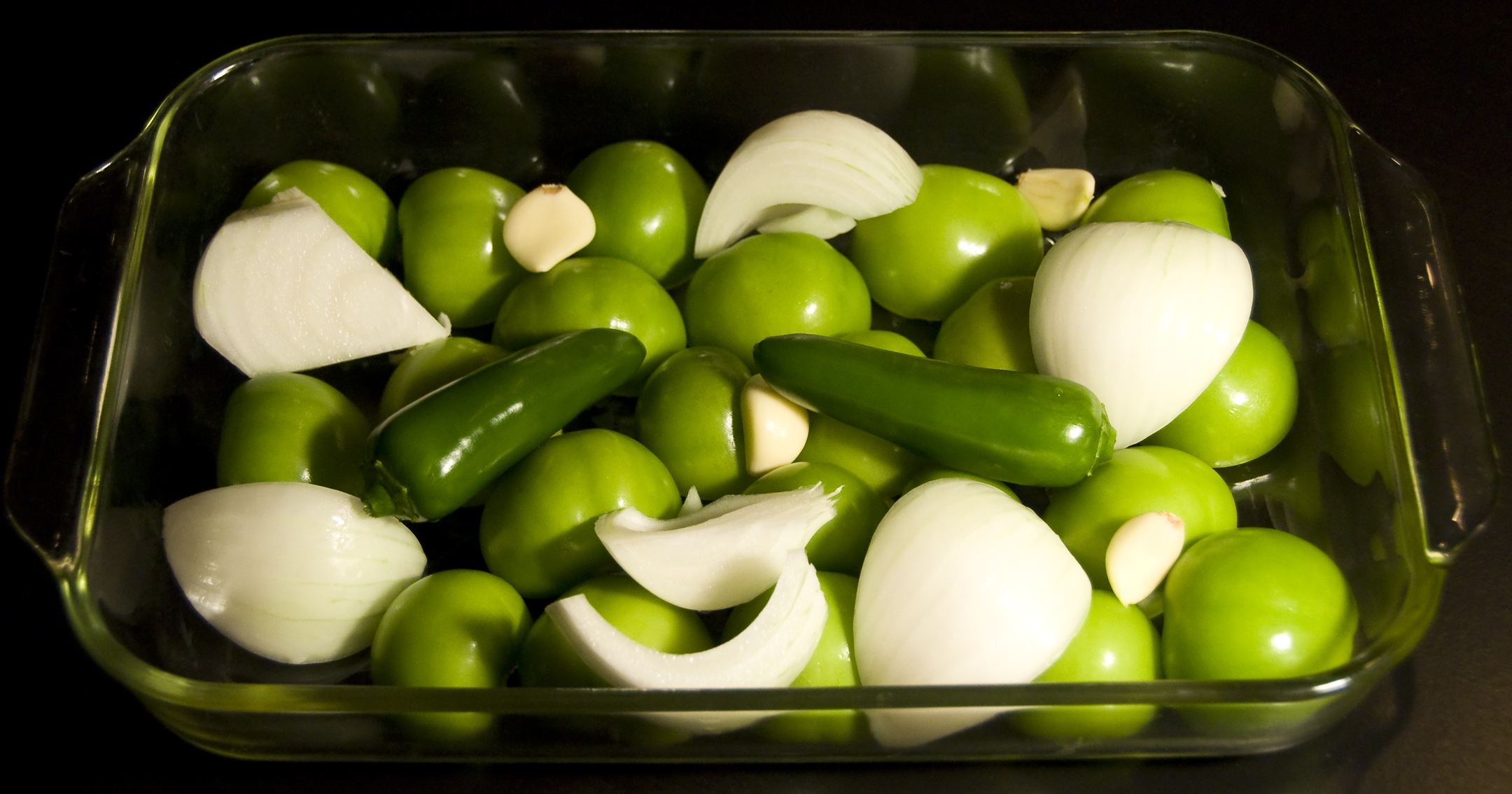 Tomatillos, onion wedges, jalapeños, and peeled garlic cloves ready for roasting on a sheet pan on a kitchen counter