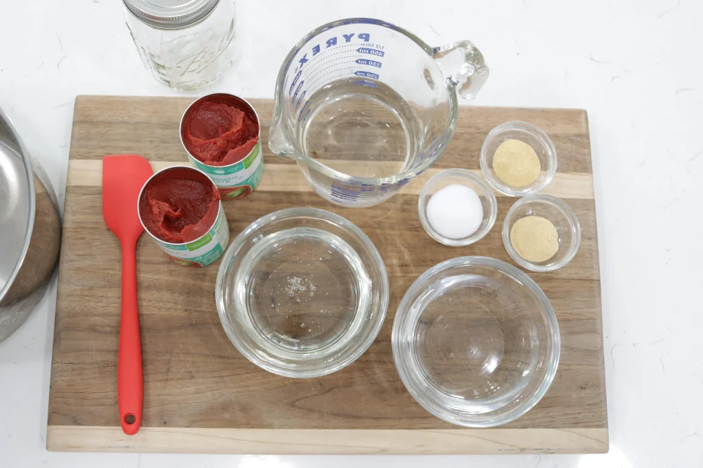 Tomato paste, apple cider vinegar, brown sugar, measuring spoons, and small bowls of spices arranged on a clean kitchen counter in warm, consistent lighting