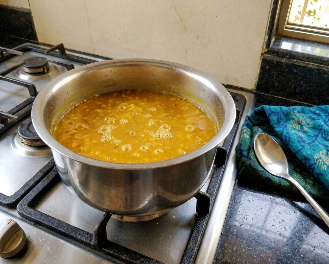 Toor dal gently simmering in a stainless steel pot on the stove with a spoon resting nearby