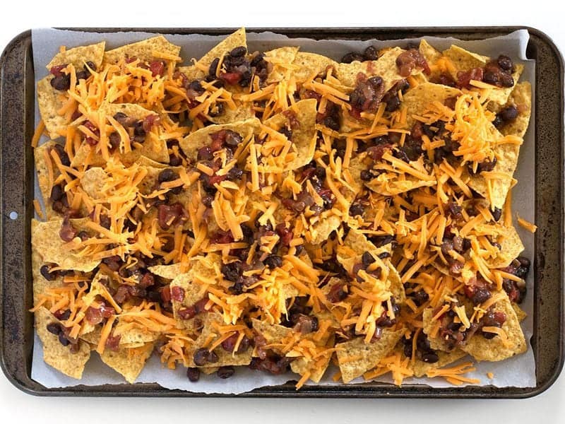 Tortilla chips, shredded cheddar and Monterey Jack, a bowl of black beans, and salsa on a counter with a lime and spices