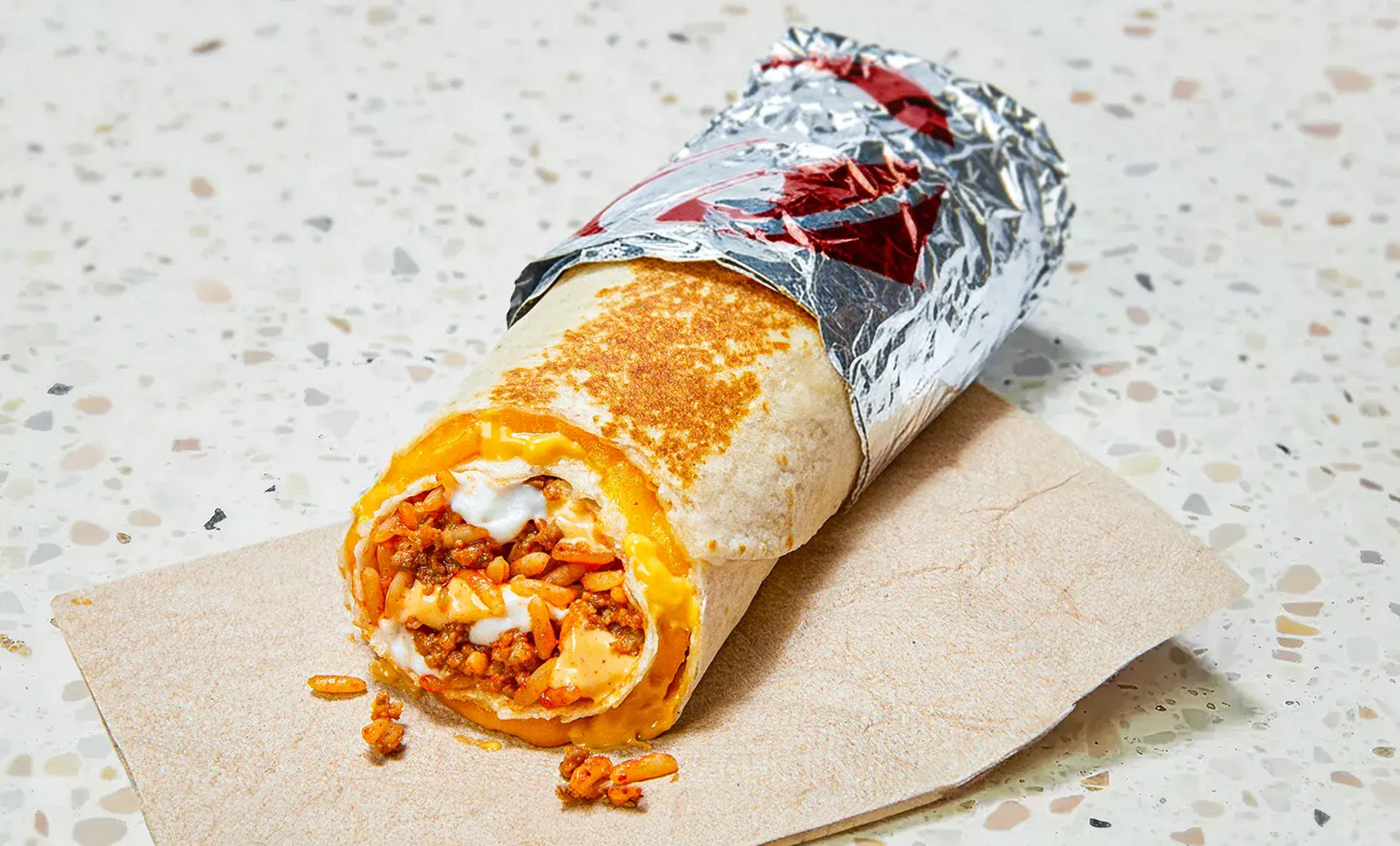 Two assembled quesaritos wrapped tightly in foil on a kitchen counter next to storage containers of rice and ground beef