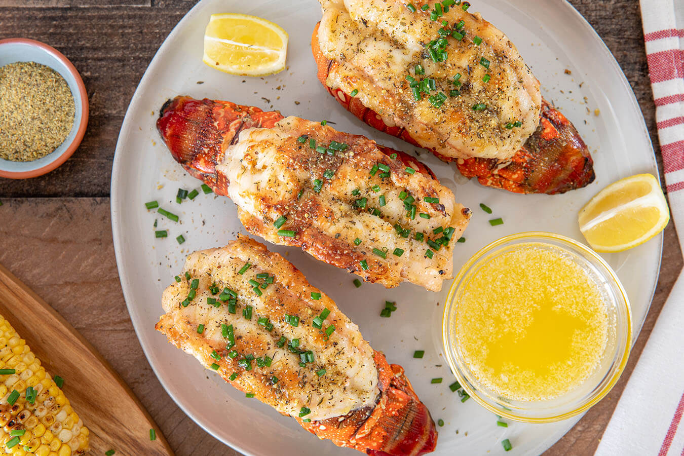 Two baked lobster tails in their shells on a sheet pan, topped with melted garlic butter and paprika, with lemon wedges on the side in a warm home kitchen