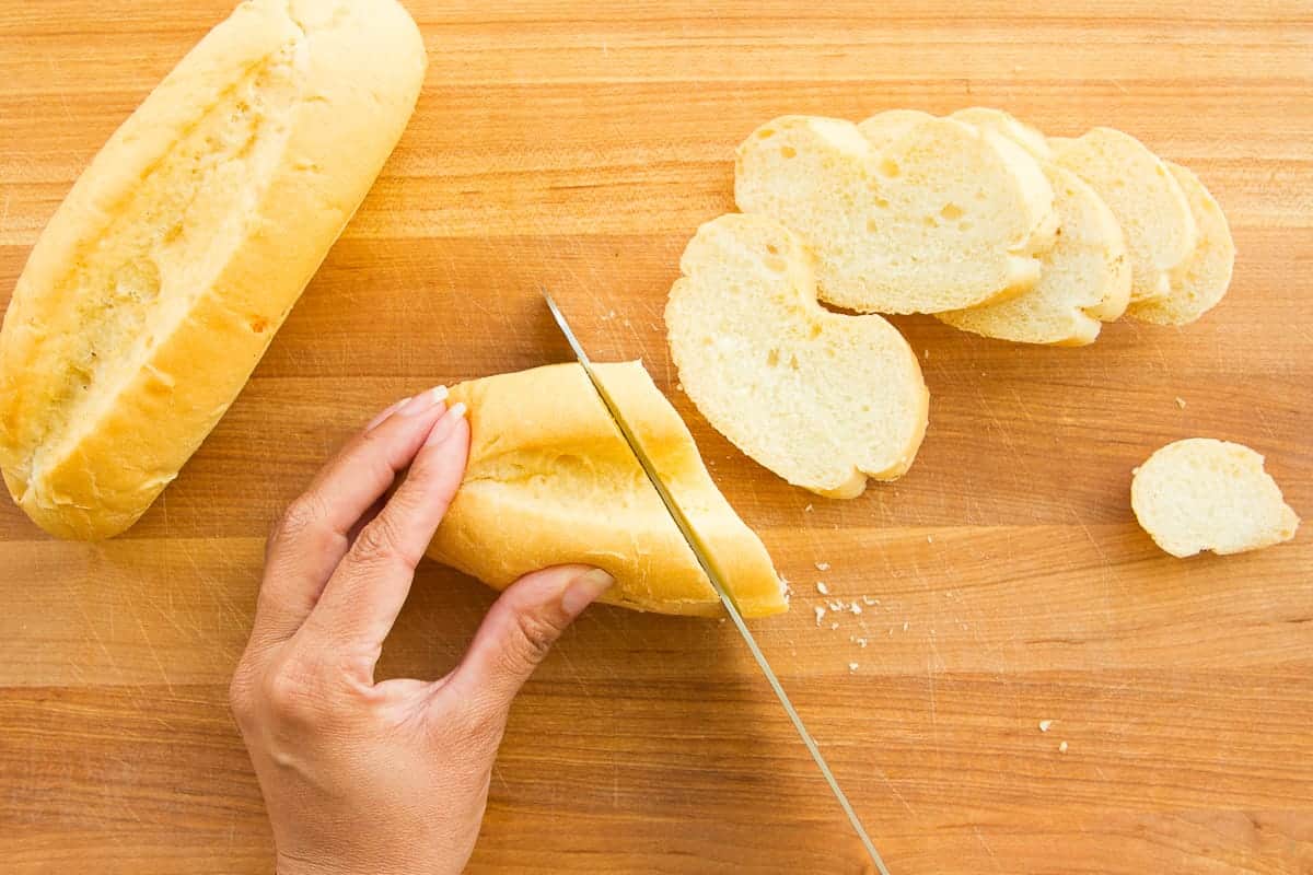 Two bolillo rolls on a wooden cutting board next to a serrated knife, ready to be sliced for tortas