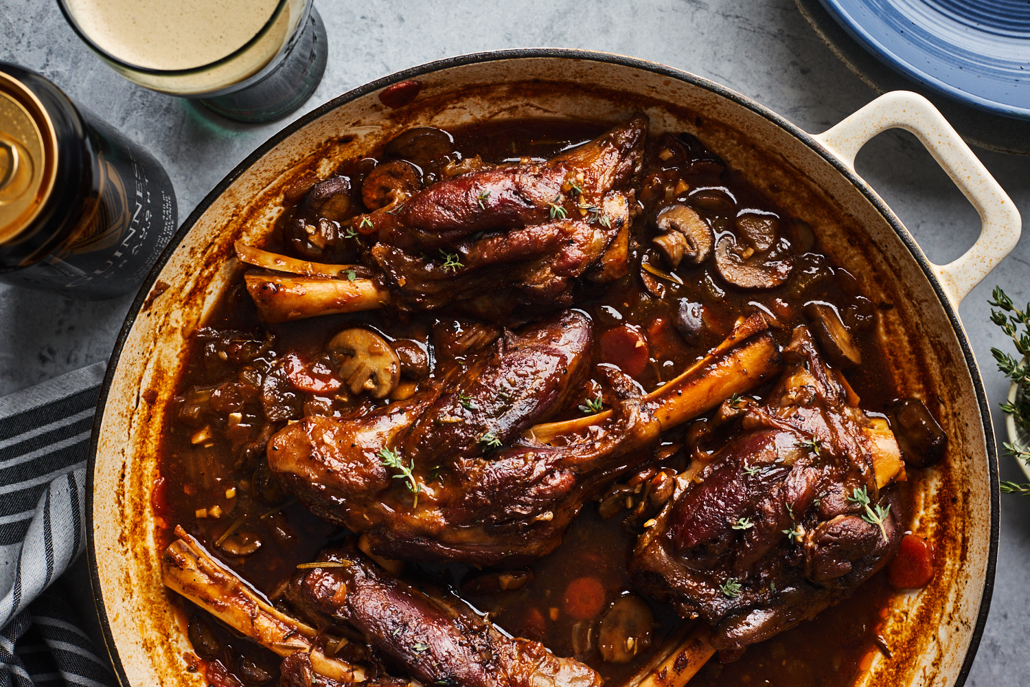 Two braised lamb shanks in a Dutch oven with a rich brown sauce, carrots, and onions, set on a warm kitchen counter with soft natural light